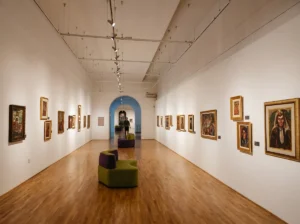 A spacious interior museum corridor with wooden parquet floors, white walls displaying framed artworks, and modern seating furniture