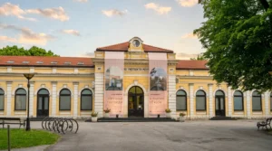 The yellow Austro-Hungarian facade of the old railway station building, home to the Museum of Contemporary Art RS, with large exhibition banners at the entrance