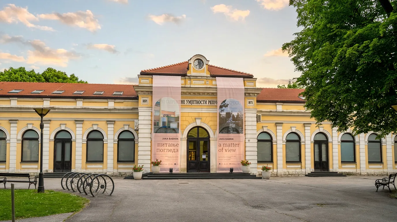 The yellow Austro-Hungarian facade of the old railway station building, home to the Museum of Contemporary Art RS, with large exhibition banners at the entrance