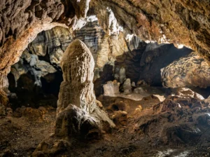 Krupni plan impresivnog, masivnog kamenog stalagmita koji se uzdiže sa tla u velikoj dvorani pećine Rastuša, sa slojevitim stijenama u pozadini