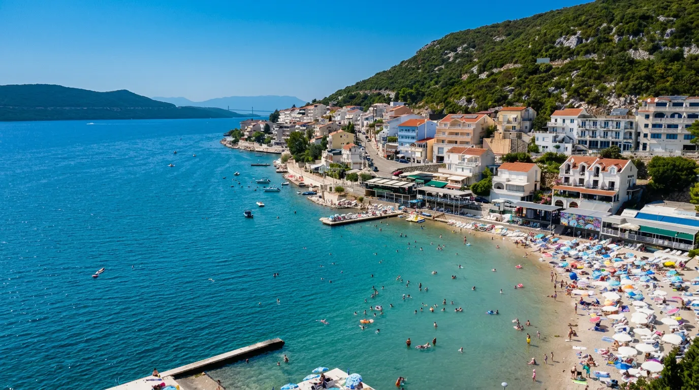 A beautiful panoramic bird's-eye view of the Neum coastline, turquoise-blue sea with swimmers, small piers, and densely built hotels and apartments along the hillside