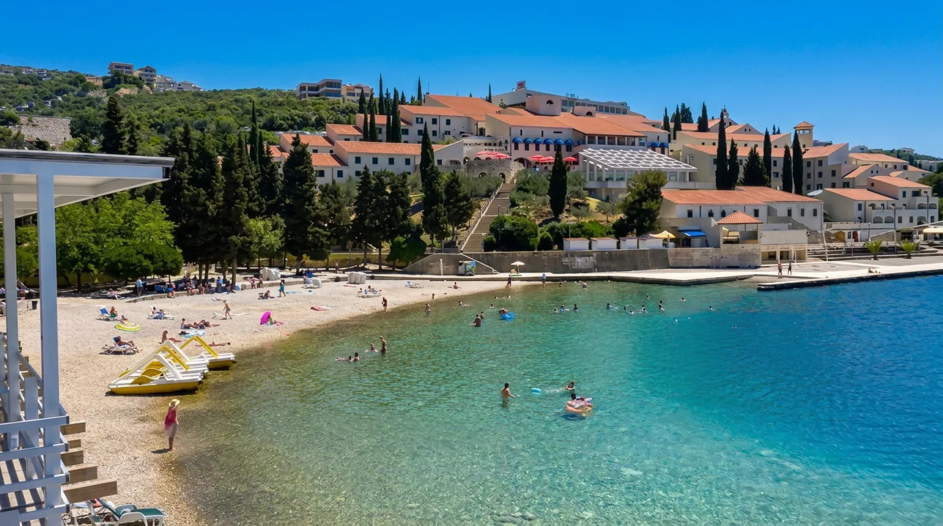 A high-angle view of Zenit Beach in Neum with clean, clear turquoise water enjoyed by swimmers, while coastal buildings and Mediterranean vegetation can be seen in the background
