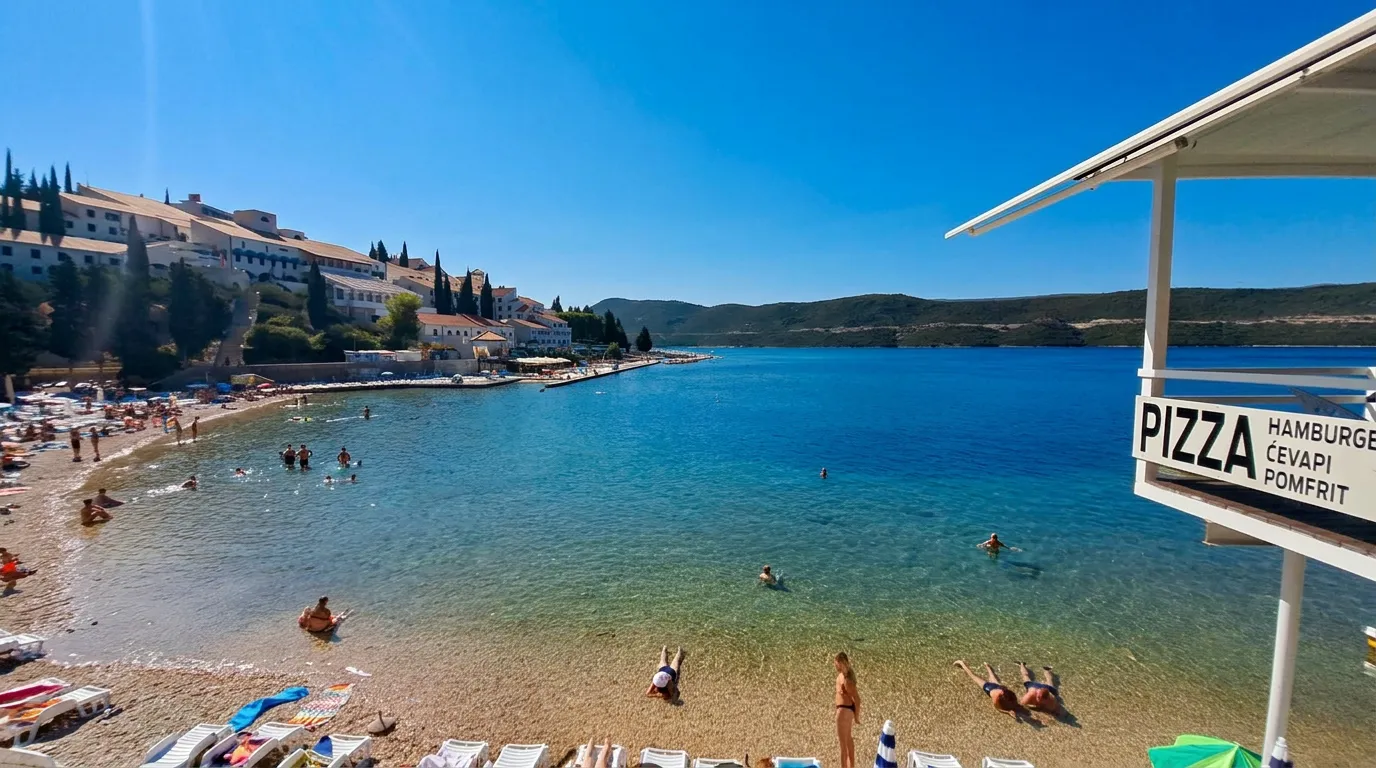 A view toward the calm and sunny Neum Bay from Zenit Beach, showing swimmers on the pebble shore and crystal-clear sea shifting from light green to deep blue