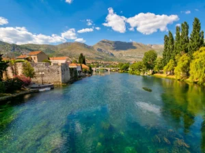 A view of the clear, turquoise water of the Trebišnjica River flowing past the stone walls of the old town in Trebinje, with a historic bridge and hills under a blue sky in the background