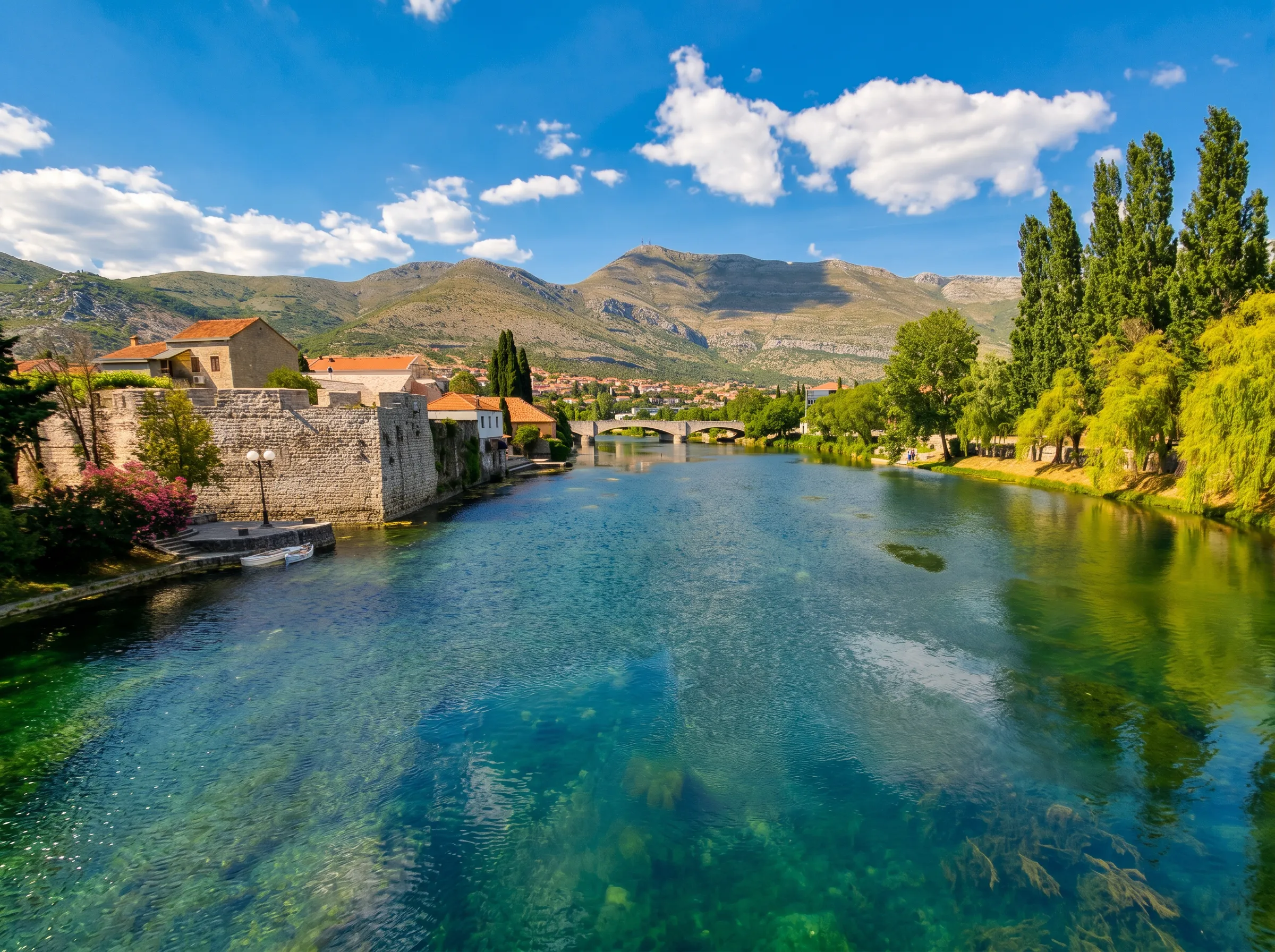A view of the clear, turquoise water of the Trebišnjica River flowing past the stone walls of the old town in Trebinje, with a historic bridge and hills under a blue sky in the background