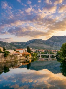 Traditional stone houses with red roofs on the bank of the Trebišnjica River, with Arslanagića Bridge in the background and the reflection of warm evening colors on the water