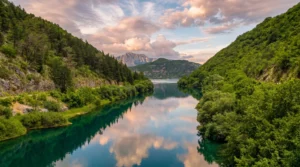 The calm, clear surface of the Trebišnjica River reflecting clouds and the surrounding hills, framed by dense green vegetation along the banks at sunset