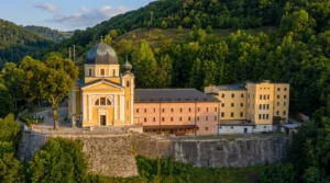 Blick auf die beeindruckende gelbe Fassade der Kirche mit großer Kuppel und das Gebäude des Franziskanerklosters in Fojnica, gelegen unterhalb eines grünen, bewaldeten Hügels