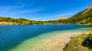 Pogled sa plitke i prozirne kamene obale na prelijepu, tirkiznu i duboko plavu vodu Šatorskog jezera, sa planinskim vrhovima u pozadini pod vedrim nebom