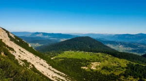 Pogled sa strme planinske padine na pošumljeno i zaobljeno brdo sa zelenim livadama u podnožju, dok se u daljini pruža širok vidik na planinske lance pod plavim nebom