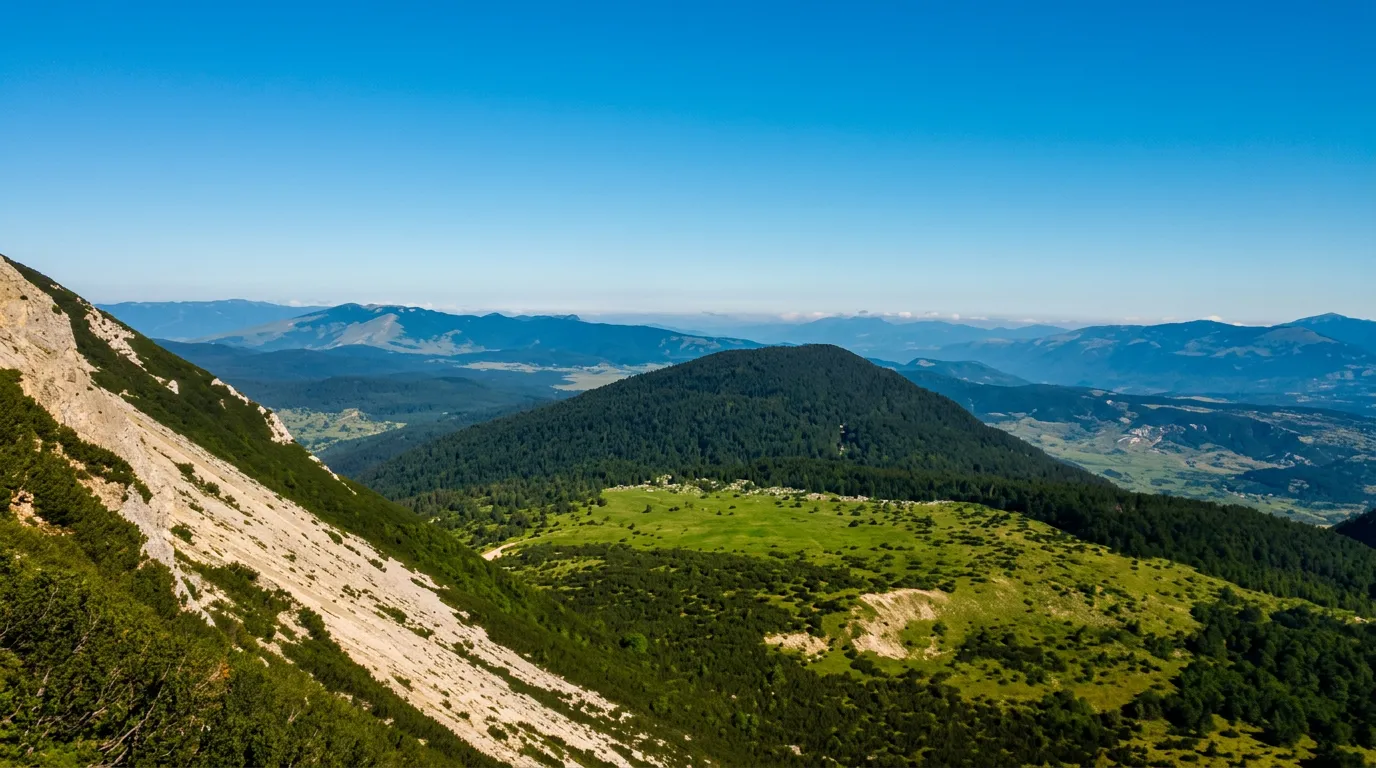 Pogled sa strme planinske padine na pošumljeno i zaobljeno brdo sa zelenim livadama u podnožju, dok se u daljini pruža širok vidik na planinske lance pod plavim nebom
