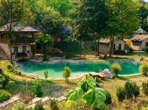 View of a clear, turquoise river pool or side channel bordered by a stone embankment, with wooden benches for resting and authentic stone buildings in a dense forest
