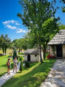 Two tourists with backpacks walk through the beautifully arranged courtyard of an ethno village with old stone houses and wooden roofs, surrounded by greenery under a blue sky