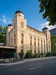 Pogled iz ugla na monumentalnu građevinu Aškenaske sinagoge sa velikim ugaonim kupolama i dekorativnom fasadom, smještenu uz gradsku ulicu pod plavim nebom