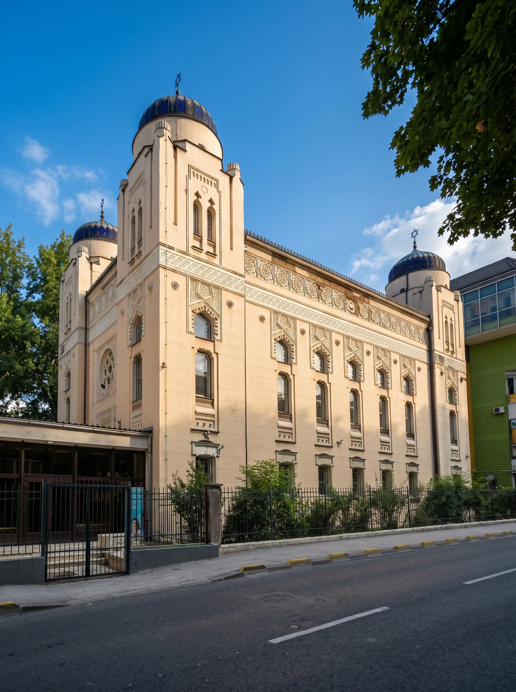 Pogled iz ugla na monumentalnu građevinu Aškenaske sinagoge sa velikim ugaonim kupolama i dekorativnom fasadom, smještenu uz gradsku ulicu pod plavim nebom