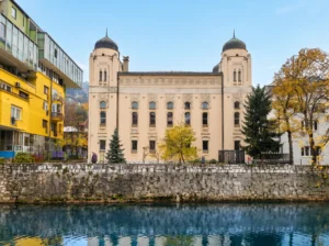 Simetrična prednja fasada Aškenaske sinagoge u Sarajevu sa dvije velike kupole, fotografisana preko bistrog toka rijeke Miljacke u kojoj se ogledaju okolne zgrade