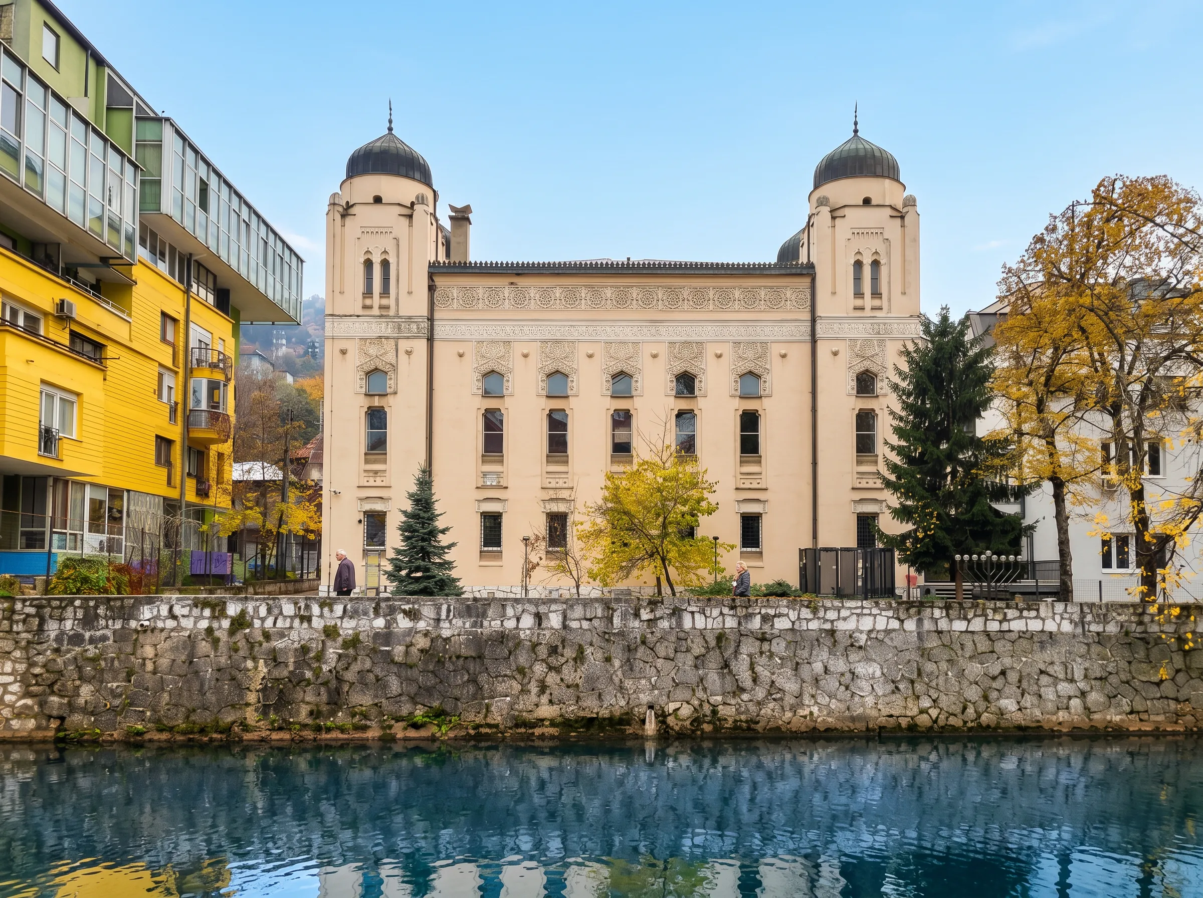 Simetrična prednja fasada Aškenaske sinagoge u Sarajevu sa dvije velike kupole, fotografisana preko bistrog toka rijeke Miljacke u kojoj se ogledaju okolne zgrade