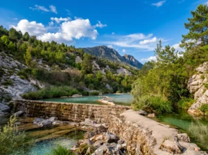 A stone travertine barrier forming a calm, clear pool on the Sušica River, surrounded by greenery with mountain peaks in the background under a blue sky and white clouds