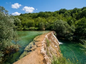View along a natural stone barrier over which water spills into a crystal-clear turquoise pool, framed by dense green forest under a sunny sky