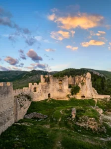 Panoramski pogled na kamnito utrdbo Stjepan-grad v Blagaju, obdano s hribi, medtem ko sta nebo in oblaki v ozadju obsijana z nežnimi toni sončnega zahoda