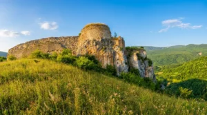 A view from the grassy base toward the remains of a massive round stone tower of the medieval Ostrovica fortress, built on a steep cliff under a clear blue sky