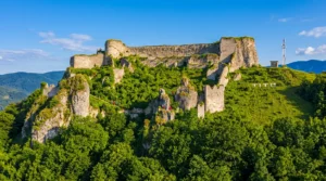 A distant view of the long defensive walls and stone towers of Ostrovica fortress rising dramatically above the dense, lush green forest at the very top of the hill