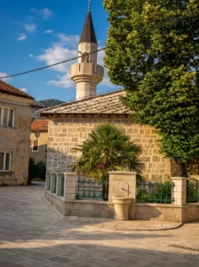 An authentic stone Osman-paša Mosque with a slender minaret, framed by a green palm tree and a decorative fountain in the courtyard inside the Old Town walls