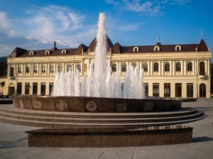 Close-up of a lavish stone fountain in the shape of a stećak, from which white jets of water spray high, with the Barok building on Trg slobode in the background under a blue sky