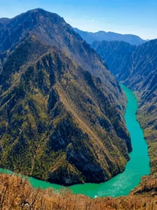 A bird's-eye view of the intensely turquoise water of the Drina River cutting through a high mountain massif with steep, rocky, and forested slopes under a clear sky.