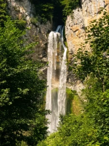 Nahaufnahme des hohen und schmalen Wasserstrahls des Wasserfalls Blihe, der über eine massive, senkrechte Felswand hinabstürzt, umgeben von grünem Laub