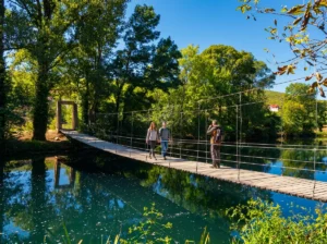 Two tourists walk across a long suspended wooden bridge with cables connecting the riverbanks, while the surrounding green trees are perfectly reflected in the calm turquoise water