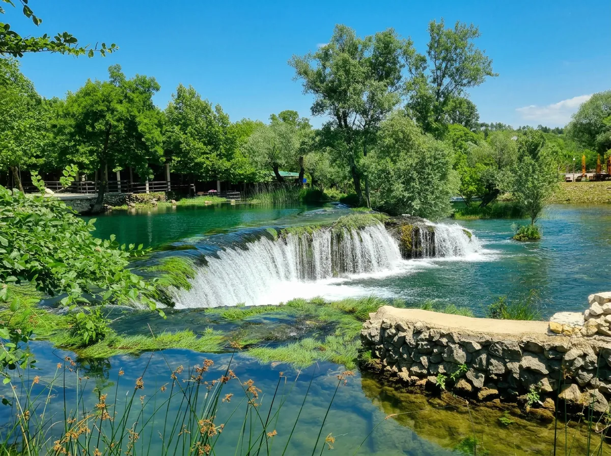 View from the riverbank of the beautiful, semicircular natural waterfall Čeveljuša, foaming and cascading into the clear green river, surrounded by lush trees and Mediterranean vegetation