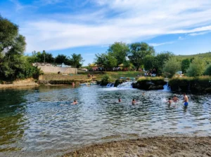 A spacious river basin and bathing area with swimmers enjoying the refreshing water in front of smaller waterfalls and rapids under a sunny, partly cloudy sky