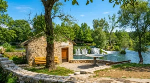 A restored old stone house with wooden doors and a landscaped courtyard with benches, located right next to the calm turquoise river flow and waterfalls in the background
