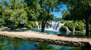 A wide series of Koćuša waterfalls flowing into the turquoise spherical riverbed, with a stone riverbank and trees in the foreground