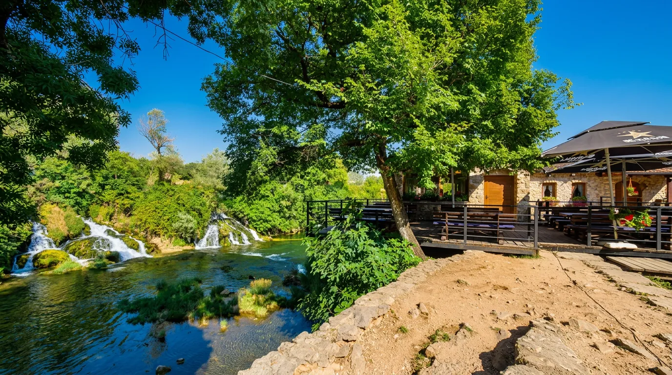 A clear river and hidden parts of Koćuša waterfalls seen from a landscaped path in the shade of a large tree