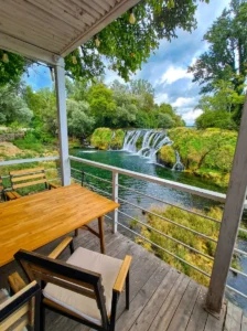 A wooden table and chairs on the terrace of a hospitality venue with a direct, open view of the wide Koćuša waterfalls