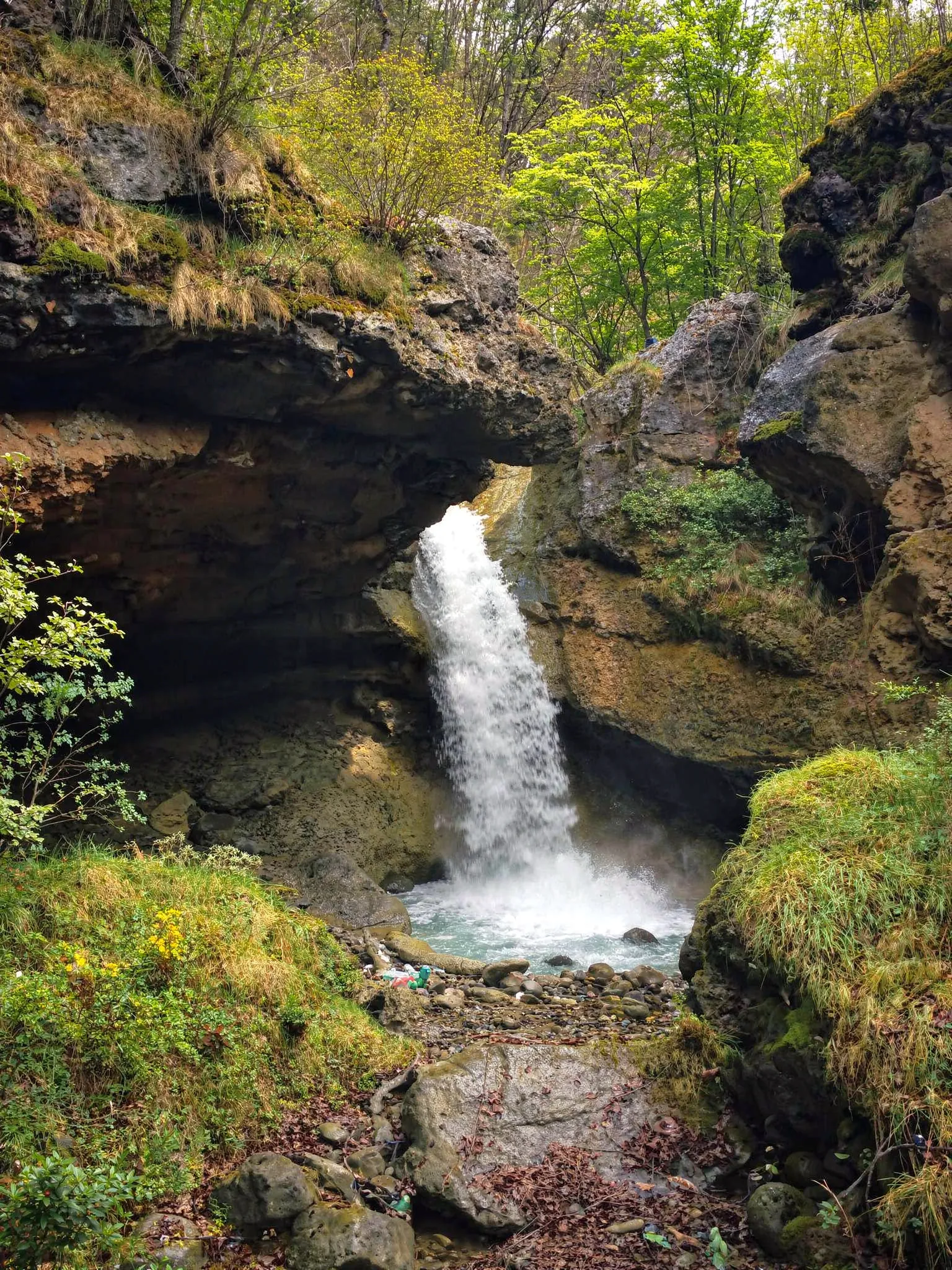 The powerful Špilice waterfall plunging through a narrow rock gap and a natural stone bridge into a small pool, surrounded by steep cliffs covered in green moss