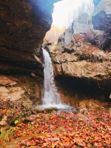 View of the foamy Špilice waterfall tumbling down a high cliff, while the ground in the foreground is covered with fallen autumn leaves in red and orange shades