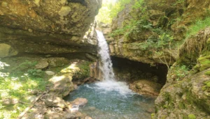 The dazzling Špilice waterfall flowing into a clear, round turquoise-green river pool hidden among massive cave rocks and lush forest greenery