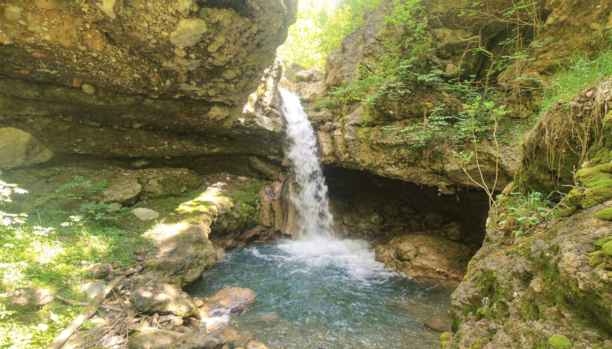 The dazzling Špilice waterfall flowing into a clear, round turquoise-green river pool hidden among massive cave rocks and lush forest greenery