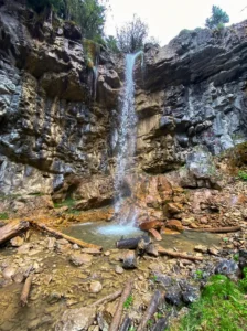 Close-up of the narrow, tall Ugrić waterfall dropping into a small foamy pool, surrounded by massive rocks and fallen wooden logs at the base