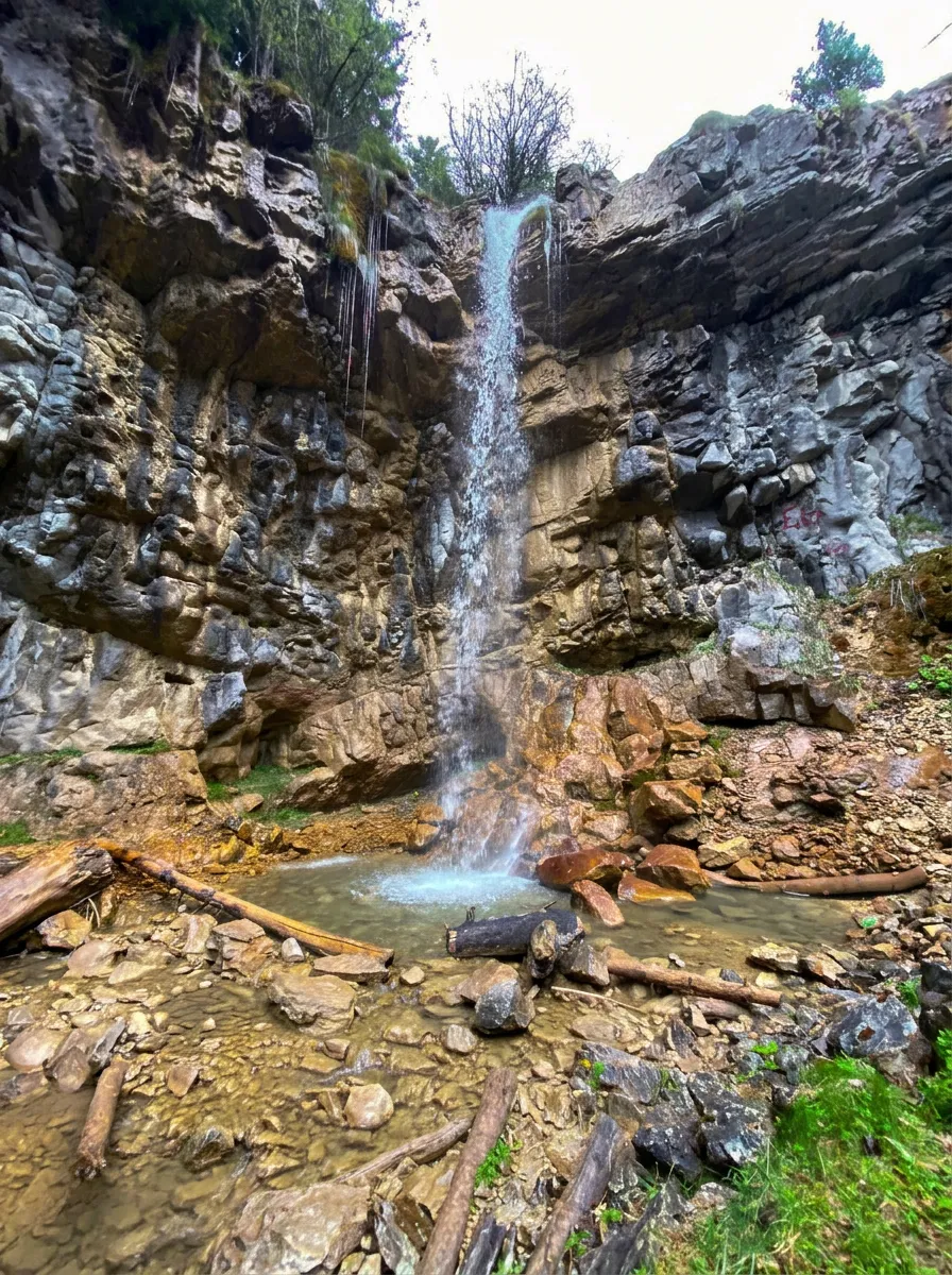 Close-up of the narrow, tall Ugrić waterfall dropping into a small foamy pool, surrounded by massive rocks and fallen wooden logs at the base