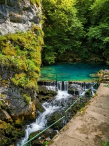 A small stone barrier at the spring of Bastašica from which the water plunges down the rocks in a fast cascade, while above it a calm natural turquoise pool is visible, surrounded by dense forest