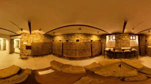 Innenraum des Ausstellungsbereichs des Heimatmuseums in Visoko mit einer halbrunden, beleuchteten Steinmauer und historischen Steinfunden und Platten, die auf dem Boden ausgestellt sind