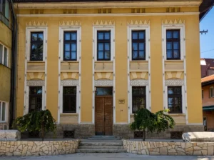 Die Vorderfassade eines historischen gelben Gebäudes mit regelmäßiger Anordnung schwarzer Fenster und hölzernen Eingangstüren, in dem sich das Heimatmuseum Visoko befindet