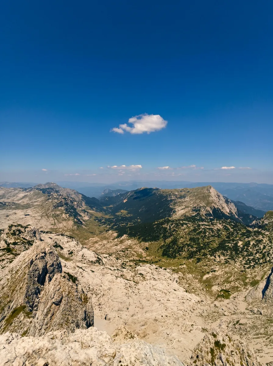 Wide panoramic view from the rocky ridge of Prenj over green valleys and distant mountain ranges under a clear blue sky with a single white cloud