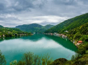 A wide panoramic view from a hill of the emerald-green winding waters of Zvorničko jezero, with houses and holiday cottages lined along the steep, forested banks