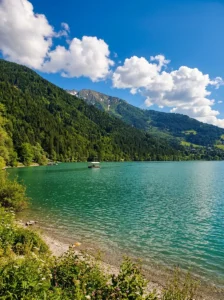 A small tourist boat or raft sailing across the vast turquoise-green water of the lake, surrounded by high mountains covered in dense coniferous and evergreen forest under a blue sky