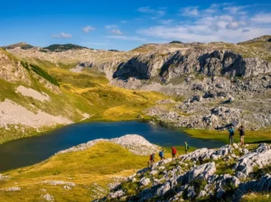 Grupa planinara sa ruksacima pješači strmim krševitim grebenom iznad duboko plavog gorskog oka — Kotlaničkog jezera, koje je okruženo masivnim kamenim liticama i žućkasto-zelenom travom.